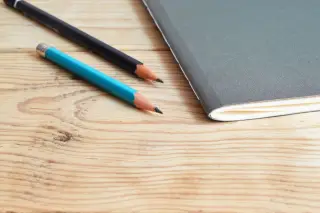 Ilustrasi: Buku catatan bersampul abu-abu dan dua pensil di atas meja kayu berpola serat alami.