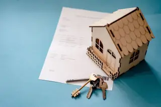 Ilustrasi: Miniatur rumah kayu, kunci, dan selembar dokumen di atas latar biru.