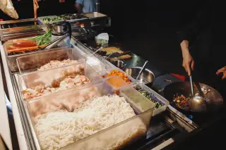 Ilustrasi: Seseorang sedang memasak di wajan dekat wadah berisi mie dan bahan makanan di gerobak kuliner jalanan.