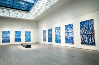 Ilustrasi: Ruang galeri seni modern yang luas dengan deretan lukisan abstrak biru di dinding putih.