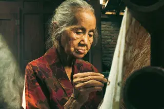 Ilustrasi: Seorang wanita lansia sedang membatik kain secara tradisional di dalam ruangan.