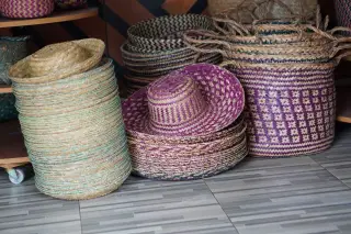 Ilustrasi: Tumpukan topi dan keranjang anyaman tradisional dengan pola warna-warni di atas lantai keramik.