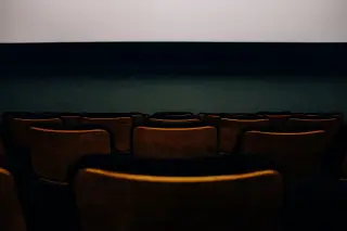 Ilustrasi: Deretan kursi teater kayu yang kosong menghadap layar putih bioskop.