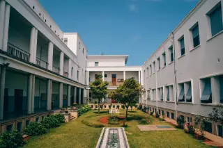 Ilustrasi: Halaman dalam gedung bertingkat putih dengan taman dan kolam kecil di bawah langit biru.