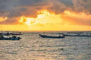 Ilustrasi: Beberapa perahu tradisional terapung di permukaan laut saat matahari terbenam dengan langit cerah berawan.