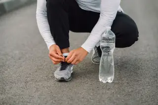 Ilustrasi: Seseorang berjongkok mengikat tali sepatu lari di samping botol air mineral di jalan aspal.