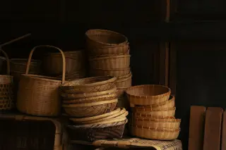 Ilustrasi: Tumpukan wadah dan keranjang anyaman bambu tradisional tersusun di dalam ruangan berlatar kayu gelap.