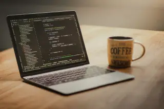 Ilustrasi: Laptop terbuka menampilkan kode pemrograman di samping cangkir kopi di atas meja kayu.