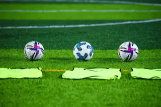 Ilustrasi: Tiga bola sepak dan rompi latihan berjejer di atas rumput lapangan hijau.