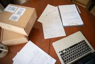 Ilustrasi: Kardus paket pengiriman, dokumen kertas, dan laptop di atas meja kerja kayu.