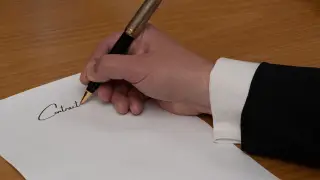 Ilustrasi: Tangan seseorang berjas menandatangani dokumen dengan pulpen di atas meja kayu.