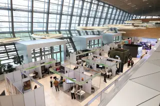 Ilustrasi: Aula pameran modern berdinding kaca luas dengan deretan stan pameran dan pengunjung.