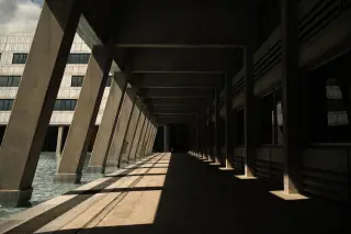 Ilustrasi: Lorong luar gedung bertiang beton tanpa orang dengan bayangan matahari.