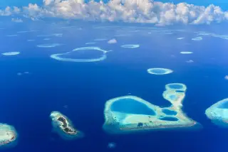 Ilustrasi: Pemandangan udara gugusan atol dan terumbu karang di laut biru di bawah langit berawan.