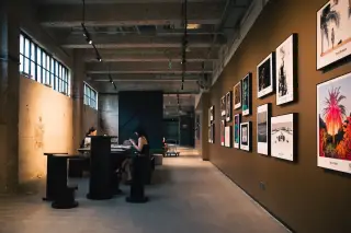 Ilustrasi: Lorong galeri seni dengan deretan foto berbingkai di dinding dan pengunjung duduk di meja.