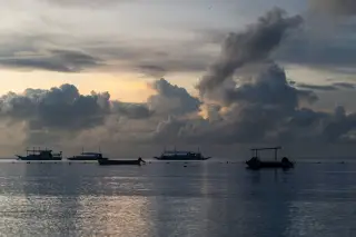 Ilustrasi: Siluet beberapa perahu terapung di permukaan laut yang tenang dengan latar awan tebal saat senja.