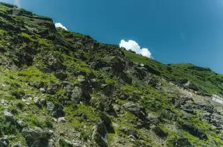 Ilustrasi: Lereng bukit berbatu yang terjal dengan tutupan rumput hijau di bawah langit biru.