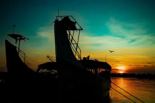 Ilustrasi: Siluet perahu nelayan bersandar di perairan tenang saat matahari terbenam dengan seekor burung melintas.