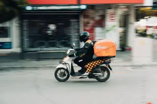 Ilustrasi: Seorang kurir bermotor dengan helm dan boks oranye melaju di jalanan kota.