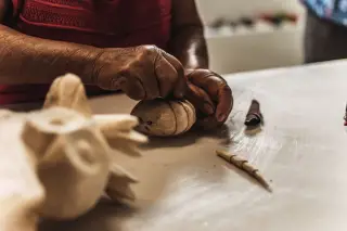 Ilustrasi: Sepasang tangan sedang mengukir dan menghaluskan kerajinan kayu di atas meja.