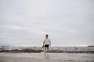 Ilustrasi: Seorang pria berjalan di tepi pantai berpasir sambil membawa kamera di bawah langit berawan.