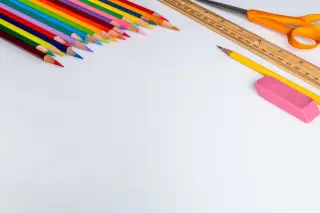 Ilustrasi: Deretan pensil warna, penggaris kayu, gunting, pensil, dan penghapus di atas latar putih.
