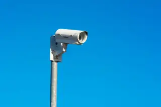 Ilustrasi: Sebuah kamera pengawas CCTV berwarna putih terpasang pada tiang dengan latar belakang langit biru cerah.