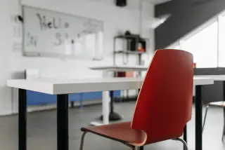 Ilustrasi: Meja dan kursi merah di ruang kelas kosong dengan papan tulis di latar belakang.