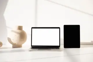 Ilustrasi: Laptop berlayar putih polos, tablet, dan vas keramik di atas meja kerja putih dengan pencahayaan lembut.