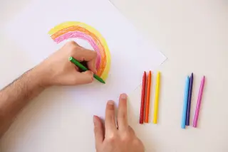 Ilustrasi: Tangan seseorang sedang mewarnai gambar pelangi dengan krayon di atas kertas putih.
