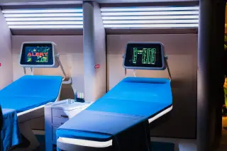 Ilustrasi: Dua ranjang medis futuristik dengan layar monitor digital di atasnya dalam ruangan berpenerangan khusus.