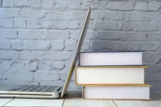 Ilustrasi: Tumpukan tiga buku tebal di samping laptop terbuka di atas meja dengan latar dinding bata.