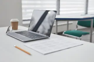 Ilustrasi: Laptop, cangkir kopi, kacamata, pensil, dan lembaran kertas di atas meja kerja putih.
