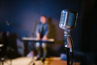 Ilustrasi: Mikrofon bergaya vintage di panggung dengan latar pemain keyboard yang buram.