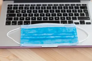 Ilustrasi: Masker medis biru diletakkan di atas papan ketik laptop yang terbuka di meja kayu.