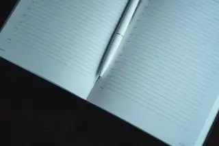 Ilustrasi: Buku catatan bergaris yang terbuka dengan pena putih di atas meja kayu gelap.