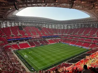 Ilustrasi: Pemandangan luas bagian dalam stadion sepak bola modern dengan tribun kursi merah dan lapangan rumput hijau.