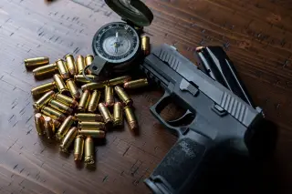 Ilustrasi: Pistol hitam, tumpukan peluru, dan sebuah kompas militer tergeletak di atas meja kayu.