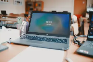 Ilustrasi: Sebuah laptop terbuka di atas meja kelas yang kosong dengan deretan meja di latar belakang.