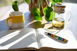 Ilustrasi: Buku catatan terbuka dengan pena, cangkir keramik, dan teko teh di atas meja kayu yang tersorot matahari.