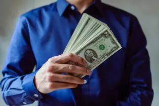 Ilustrasi: Seseorang berkemeja biru memegang segepok uang kertas dolar AS.