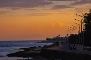 Ilustrasi: Pengendara motor melintasi jalan pesisir pantai saat senja dengan deretan tiang lampu.