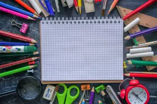 Ilustrasi: Buku catatan bergaris kotak dikelilingi berbagai alat tulis dan perlengkapan sekolah.
