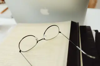 Ilustrasi: Kacamata berbingkai bulat tergeletak di atas buku terbuka di dekat laptop.