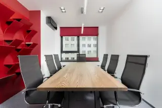 Ilustrasi: Meja rapat kayu panjang dikelilingi kursi kerja hitam di ruang kantor modern dengan rak merah.