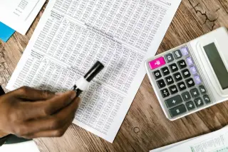 Ilustrasi: Tangan memegang pena di atas lembaran dokumen angka finansial di dekat kalkulator.