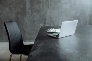 Ilustrasi: Sebuah laptop perak terbuka di atas meja kerja abu-abu dengan kursi hitam.