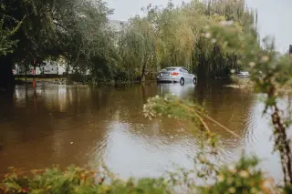 Ilustrasi: Sebuah mobil sedan perak terendam genangan air banjir di jalanan yang dikelilingi pepohonan.