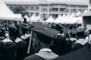 Ilustrasi: Kerumunan wisudawan bertoga dari arah belakang dalam suasana upacara wisuda luar ruangan.