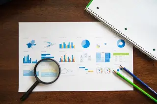 Ilustrasi: Kaca pembesar, pensil, dan buku catatan di samping lembaran grafik di atas meja kayu.